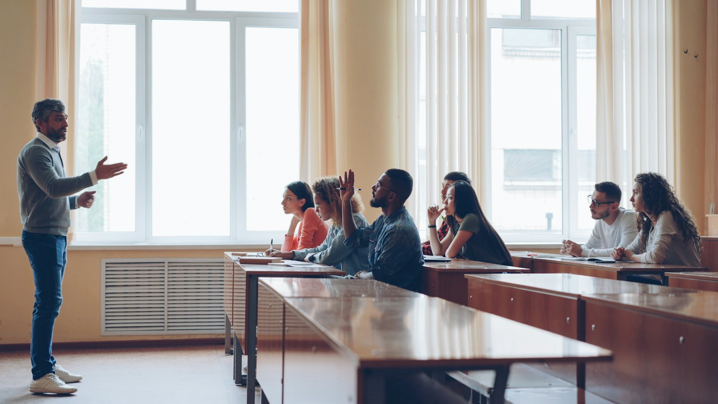 Teacher gesturing at students in a bright classroom.